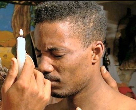 Man holding candle to face of other man as he holds his eyes closed