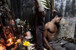 Man asleep next to Palo Mayombe altar and another man standing next to him, touching his head